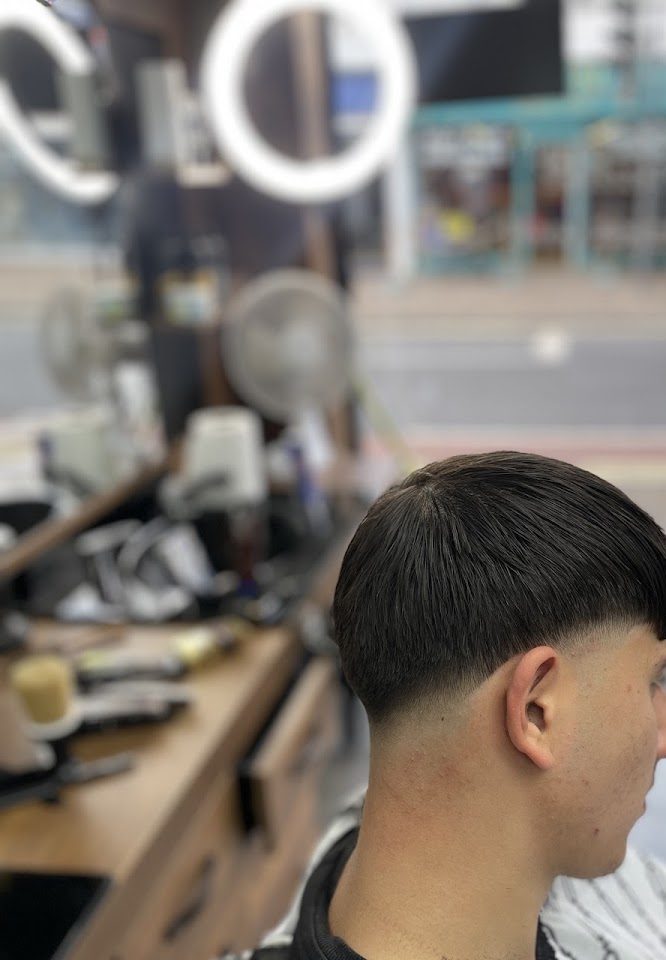 A customer in the chair from behind, hair freshly cut, with the barber's counter, clippers and ring light in the background and the High Road through the window